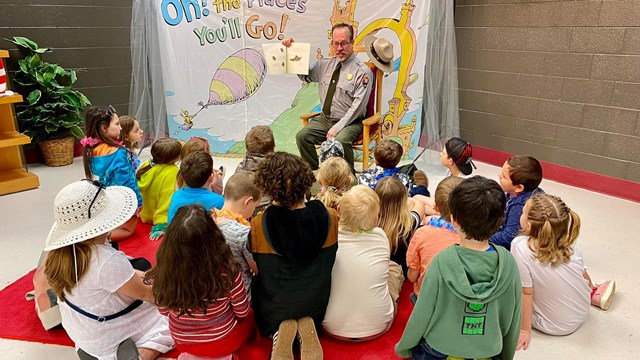A seated uniformed man holds high an open book, reading to 20 kids seated on the floor in front.
