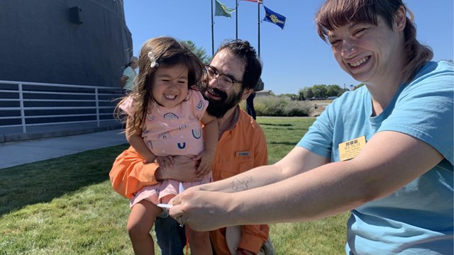 A smiling man holds a smiling child while a young woman holds onto a paper crane with her arms outst