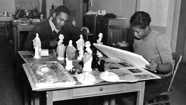 A black and white photo of two people at table covered in figurines and plaster models.
