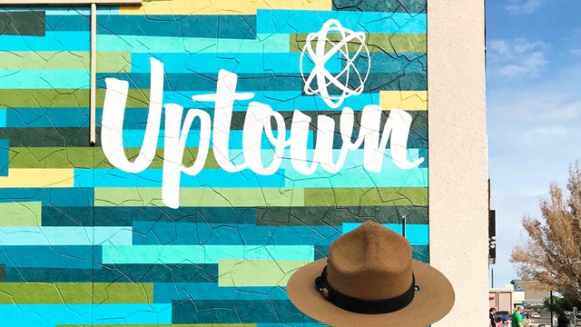 A color photo of a woman in a flat hat standing before a building with the word "Uptown."