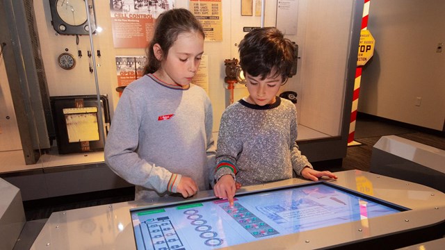 Two children look at a screen built into a table. An exhibit is behind them. 