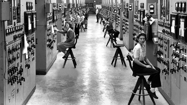 Black and white photo of women sitting in front of walls full of dials and knobs. 