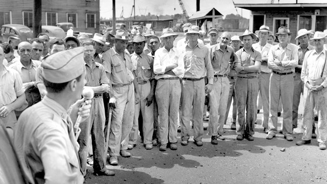 Black and white photo of a large group of men in light colored hats, shirts, and pants. 