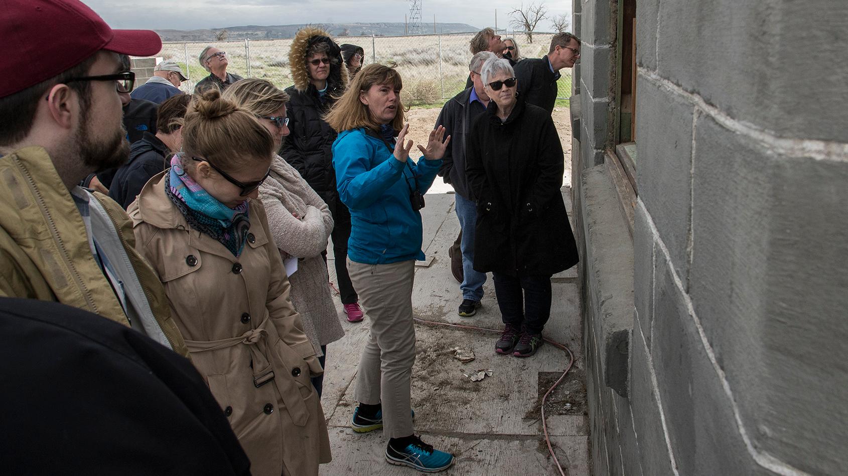 A group of people stand around a woman who is gesturing towards the gray wall of a building.