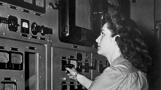 A black and white photo of a woman in front of a wall of dials.