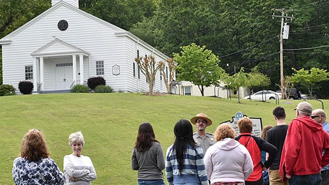 A uniformed park ranger guides a group of visitors in front of a grassy field below a white church.