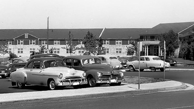 Black & white photo of classic cars in parking lot in front of long two-story building.