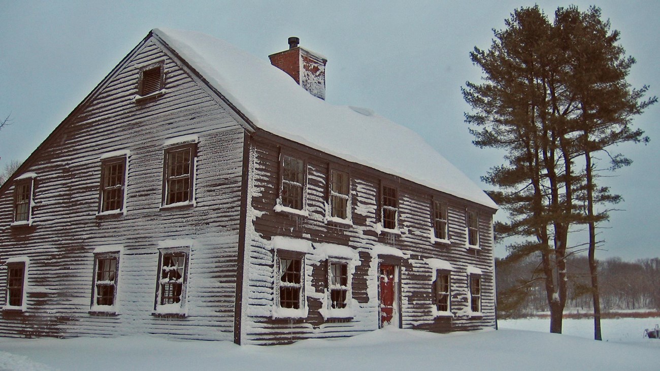 Meriam House covered in snow with a pine tree in the background and a gray sky. 