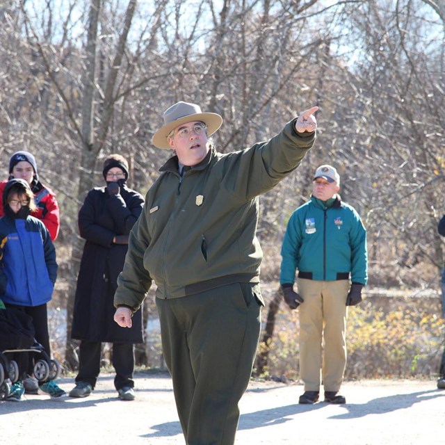 A National Park Ranger surrounded by visitors points off camera while speaking