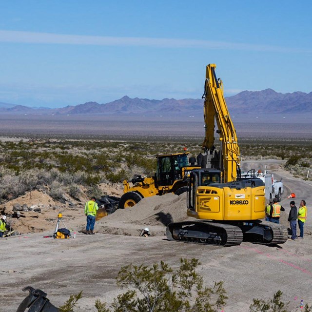 A large yellow excavator and loader overshadows workers in a shallow trench on a desert roadway