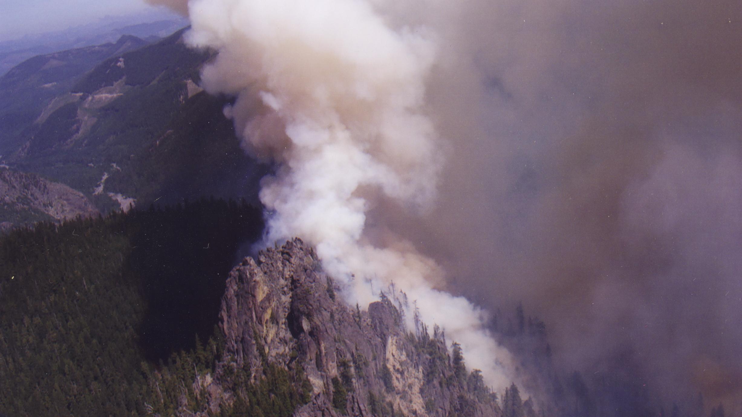 A plume of wildfire smoke rises from behind a forested mountain ridge.