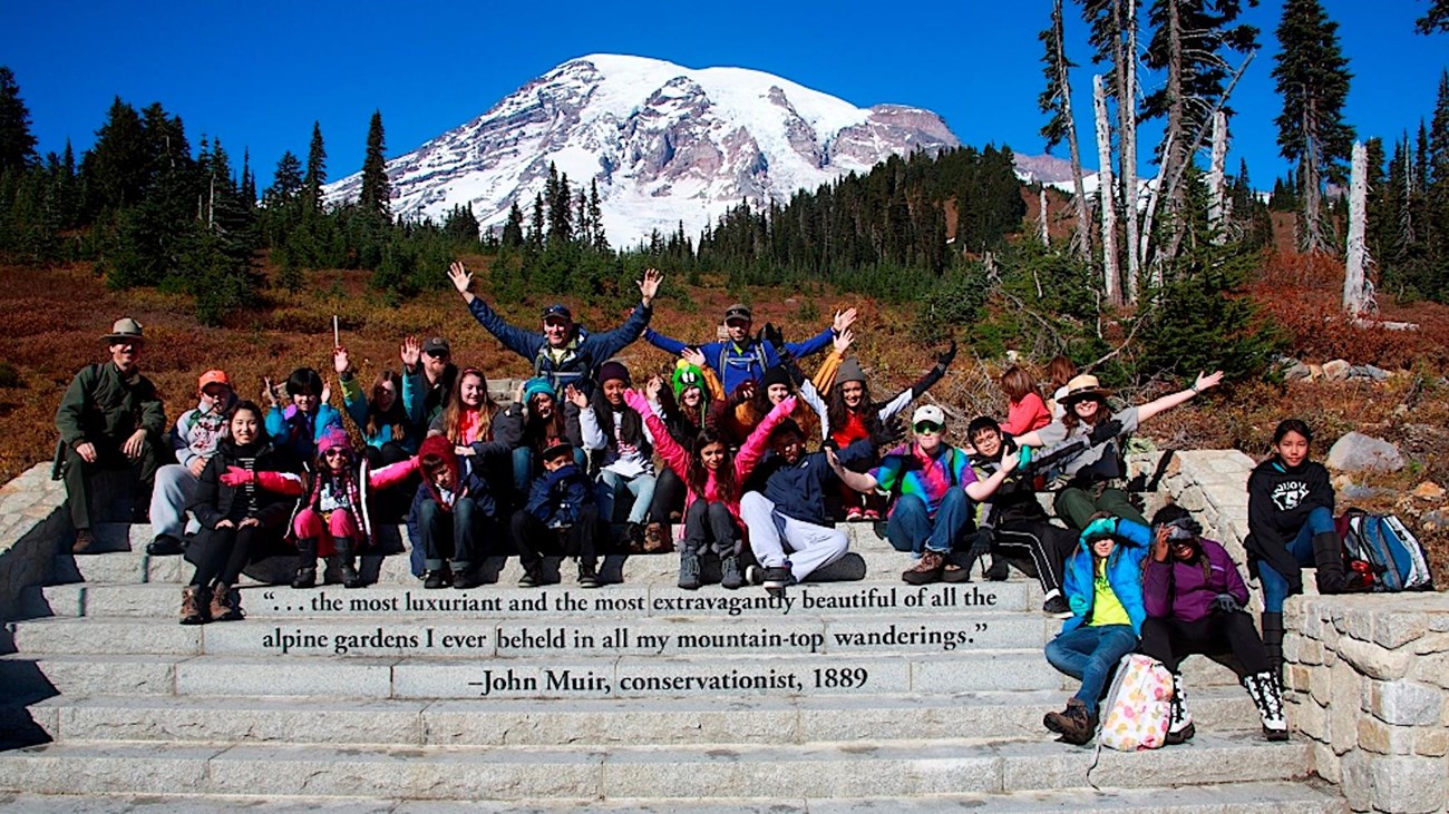 A group of students and teachers sit on stone steps in front of a view of Mount Rainier. 