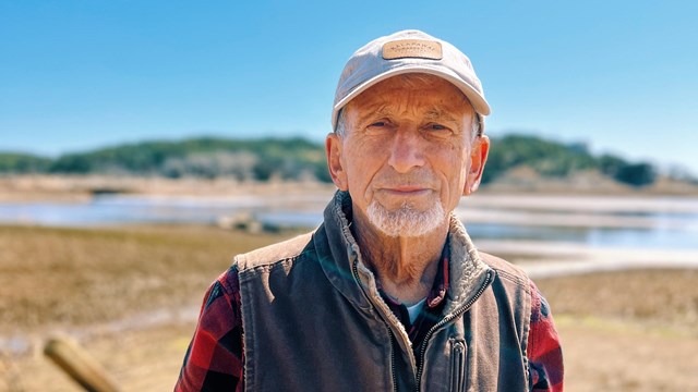 Portrait of John Portnoy. He is wearing a hat and vest, standing in front of the Herring River.