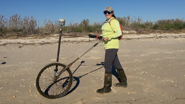 Woman stands with GPS equipment on wheels for shoreline monitoring on the beach.