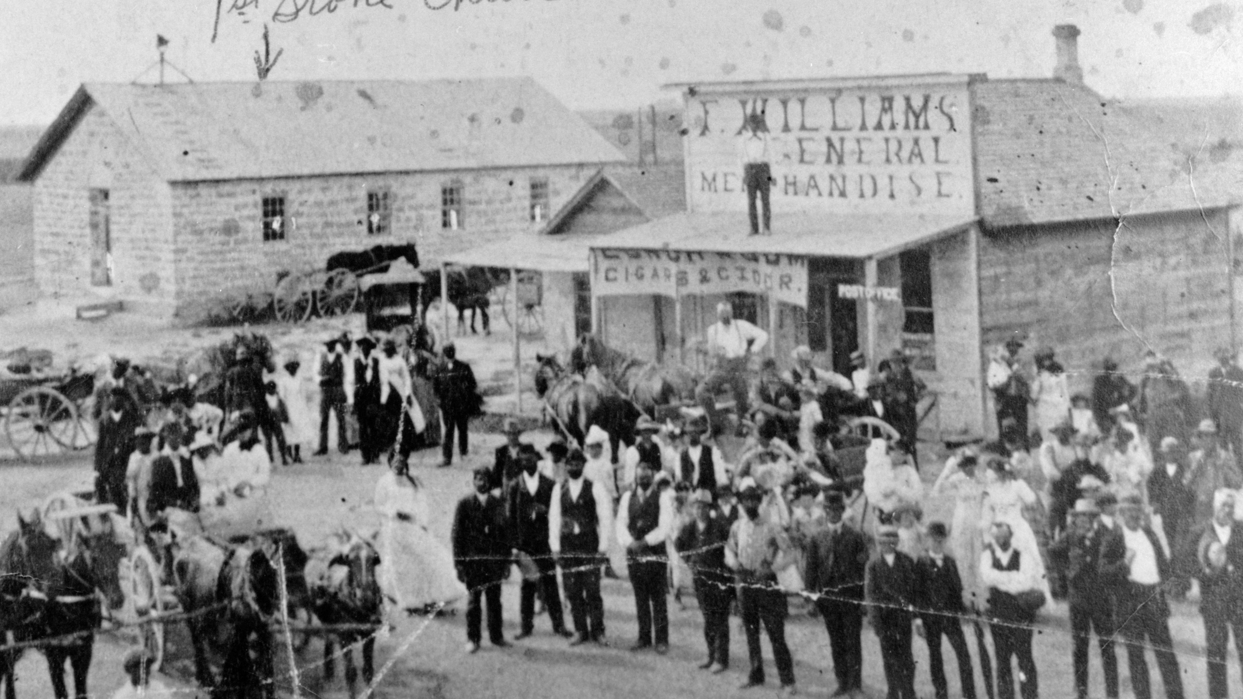 Black and white photo of many well-dressed people standing in front of frame and stone buildings.