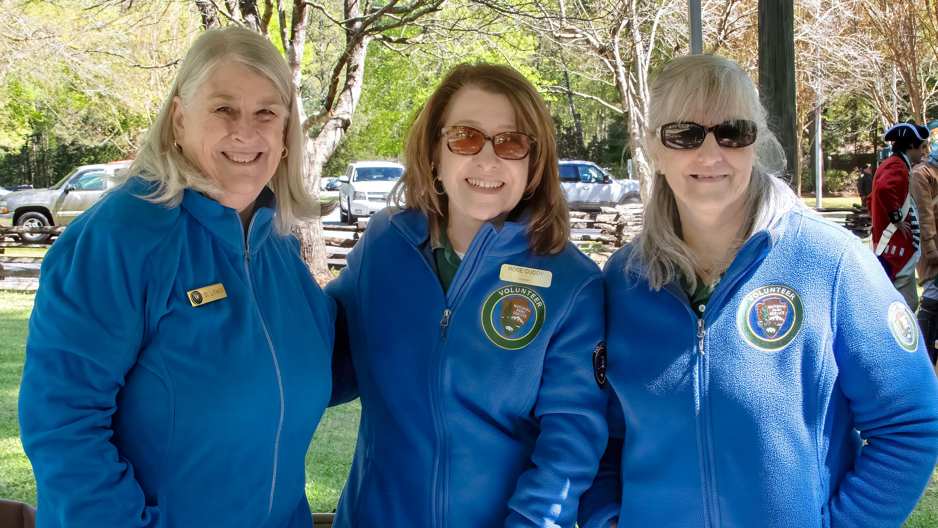 Three women in blue volunteer jackets stand at a children's table. 
