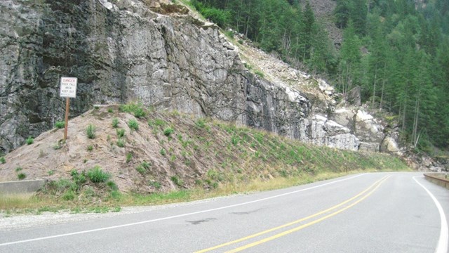 A two-lane road curves along a steep, rocky embankment.