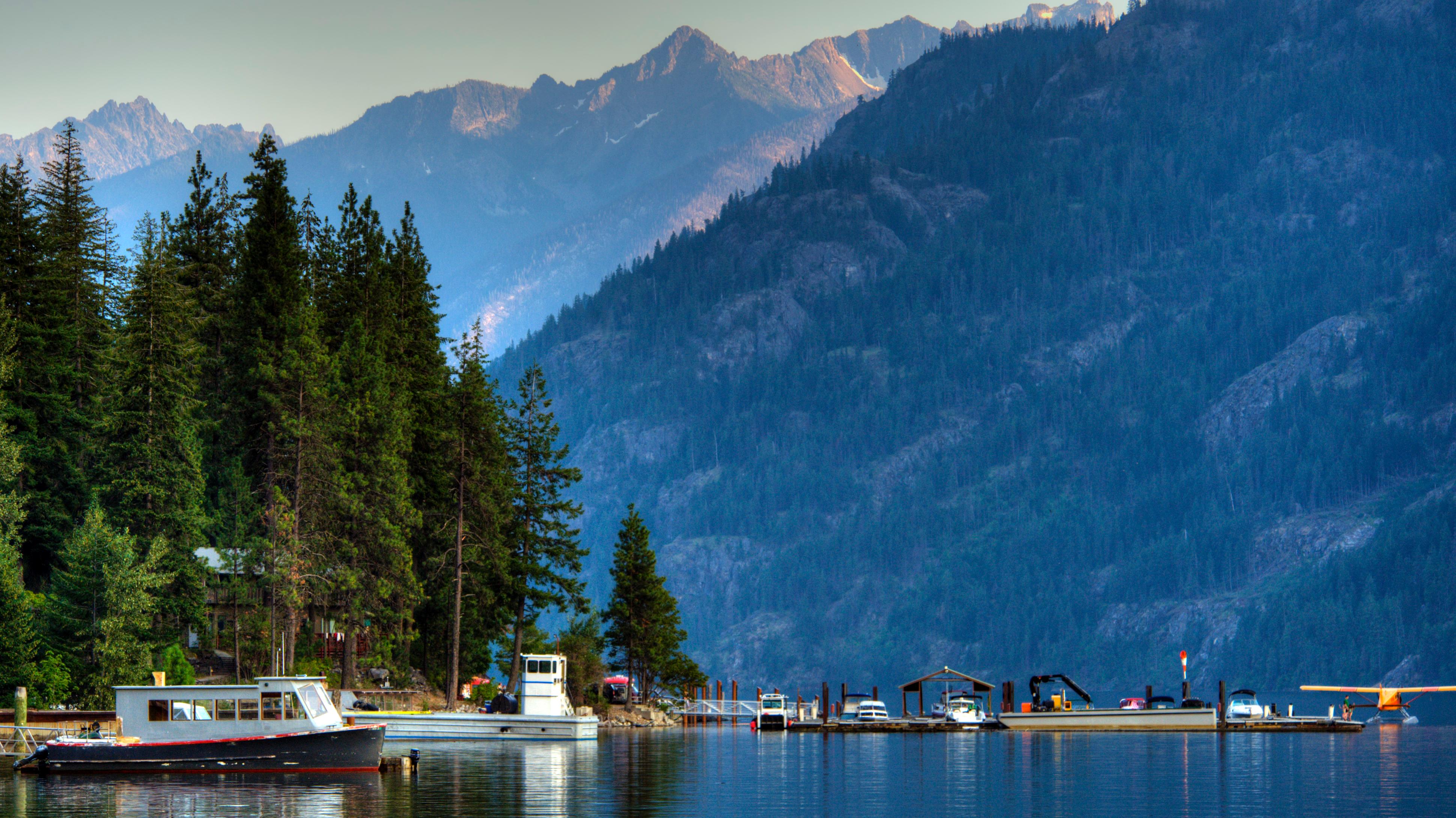 Boats and float plane docked along the shores of Lake Chelan with the mountains rising above. 