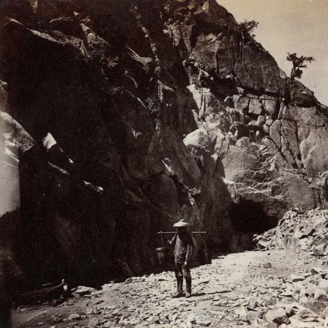 Historic black and white photo of a Chinese immigrant carrying supplies in a rocky area