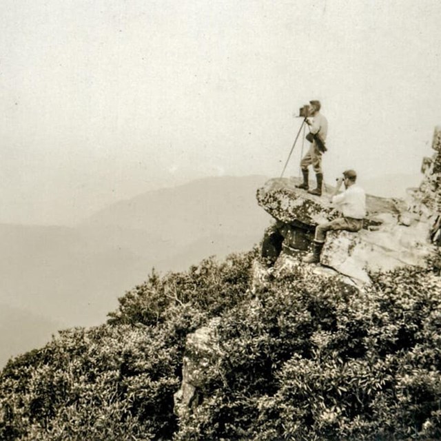 Historic image of two people standing on a rocky outcrop on a forested mountain