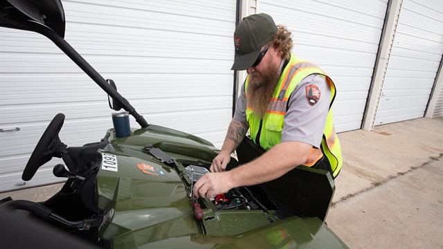 A person in a reflective vest works on the engine of a small vehicle. 