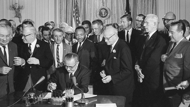 President Lyndon Johnson signs a document with Martin Luther King Jr and others standing behind him