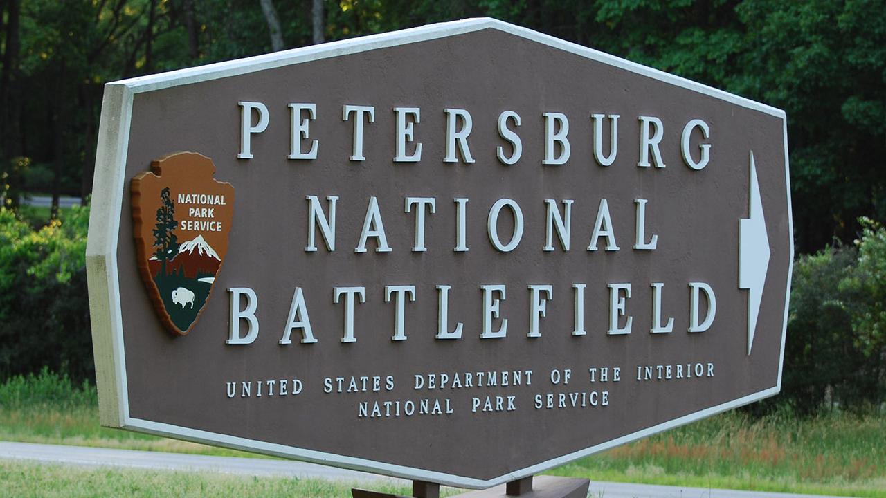 Brown sign with white text reading Petersburg National Battlefield. A NPS arrowhead is on the left.