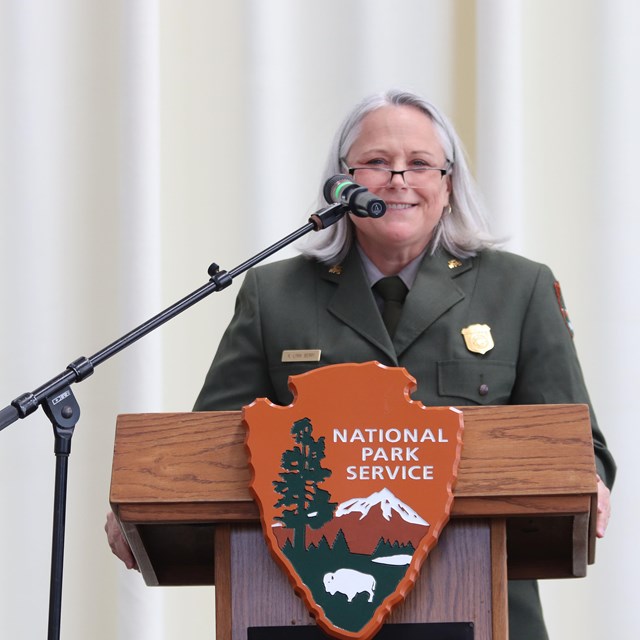 An older female park ranger smiles while standing behind a podium and microphone. 