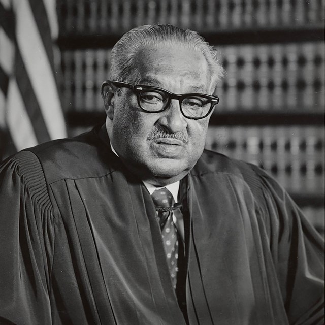 An older African American male in judicial robe with a flag and books behind him.