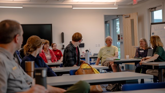 A group of people sitting look at a person speaking from their chair in the back of the room.