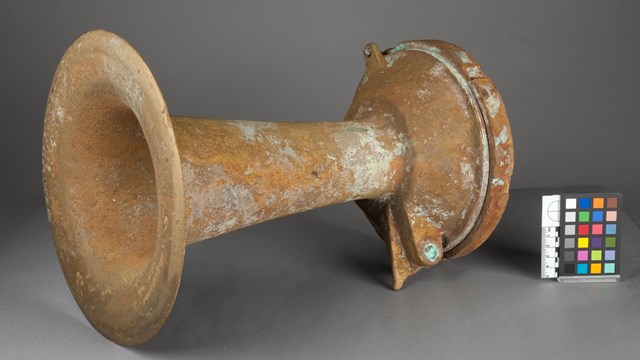 A photograph of a yellow, aged, fog horn against a gray background.