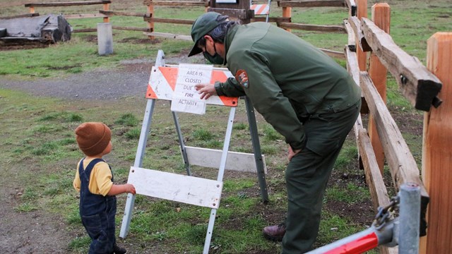 A park ranger points to a sign saying this area is closed due to fire danger, a small boy looks on.