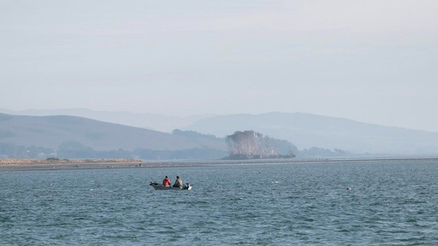 Two people in a small boat on blue waters in front of hazy, treed ridgelines. 