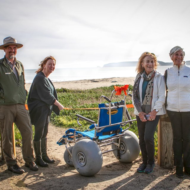 Four people stand smiling around a blue wheelchair on a coastal pathway leading to the ocean.