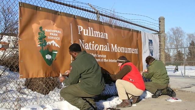 Photo of three national park staff members attaching a monument banner to a chain link fence.