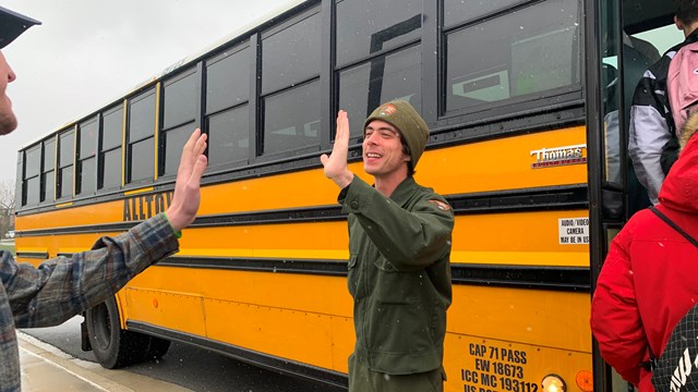 A ranger holds out his hand for a high five as students load onto a yellow school bus behind him.