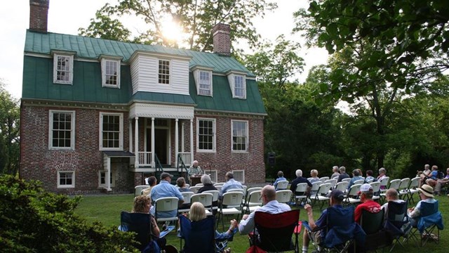 A group of people seated listening to a speaker in front of an historic house.