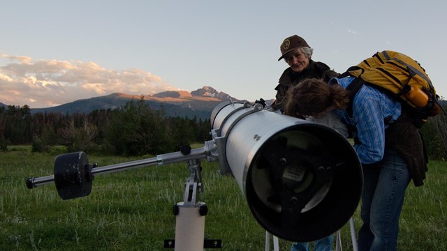 An NPS volunteer is standing by a telescope. A visitor is looking in the telescope