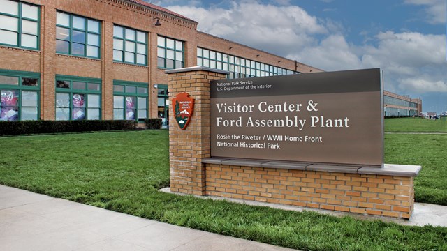 A brick sign says "Ford Assembly Plant and Visitor Center". Two-story brick building behind it. 