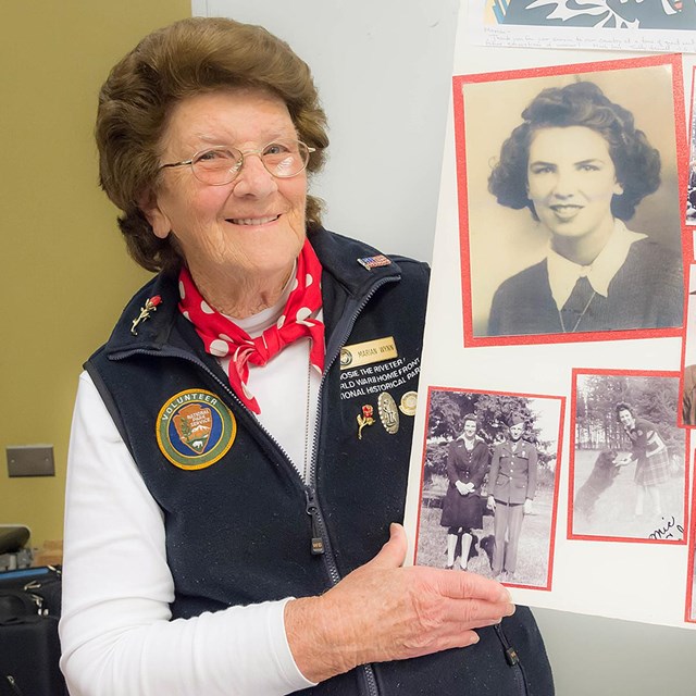 An older woman in an NPS volunteer uniform shows a homemade board with photos. 