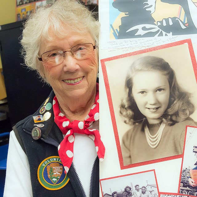 An older woman in an NPS volunteer uniform shows a self-made board with historic photos. 
