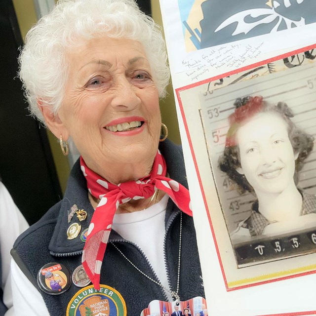An older women in an NPS volunteer uniform holds a self-made board with historic photos. 