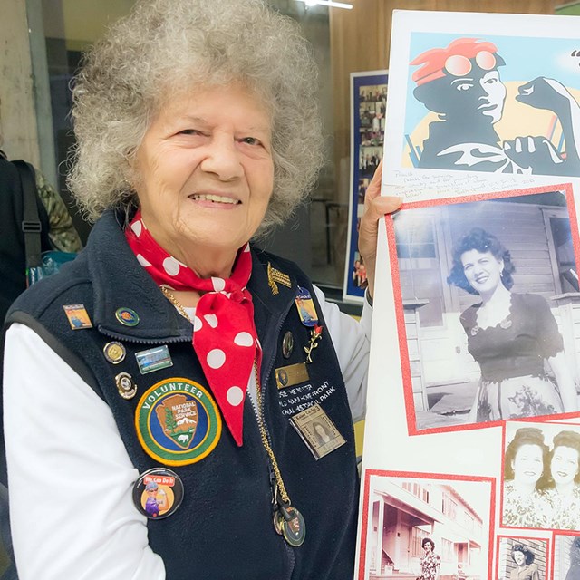 An older woman in a volunteer uniform holds a self-made sign with historic photos. 