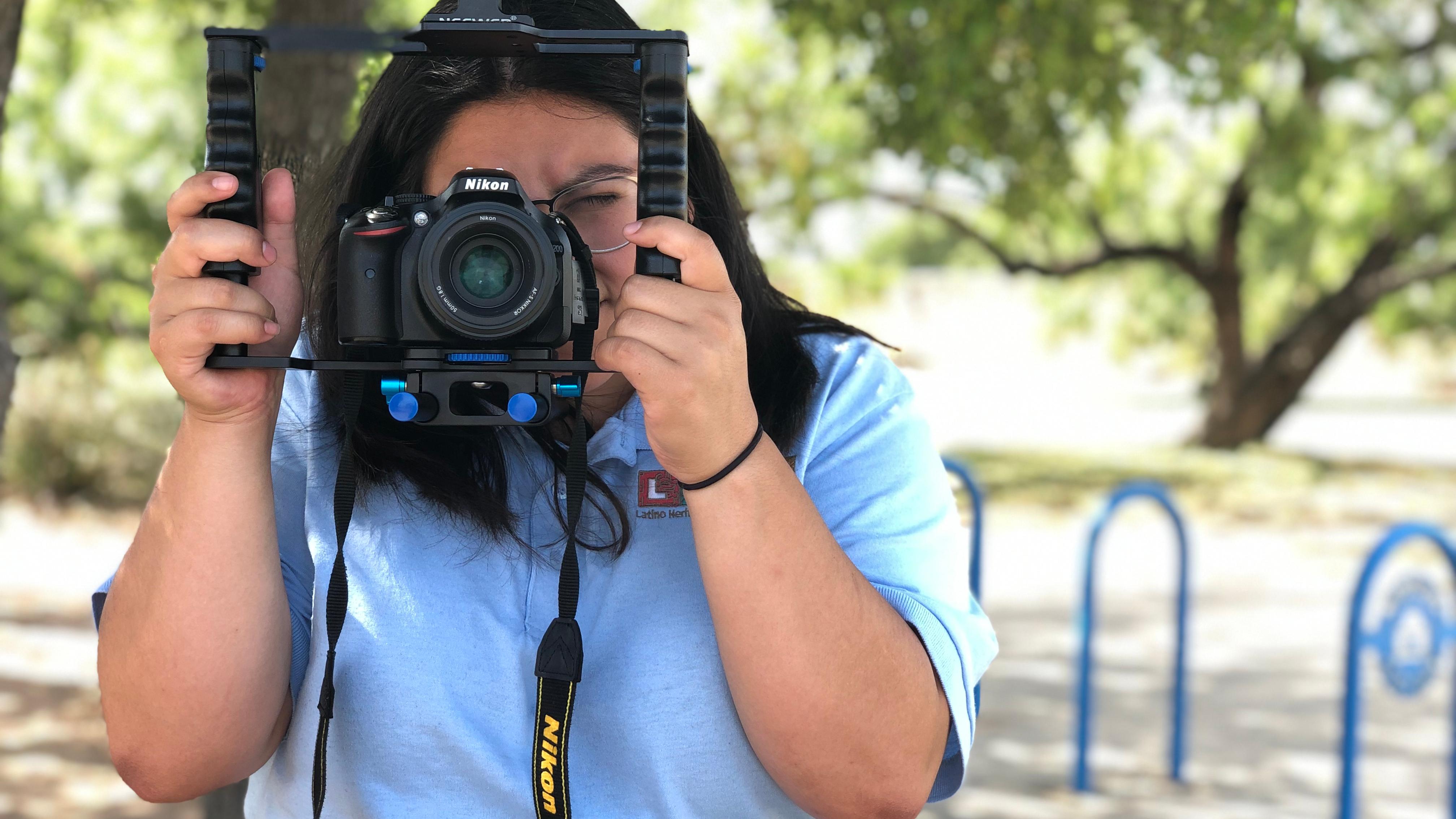 Woman holding a camera up to her face with trees behind.