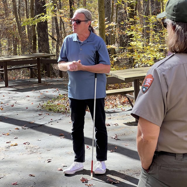 A man stands wearing sunglasses and holding a white-tipped cane. A park ranger is on the right.