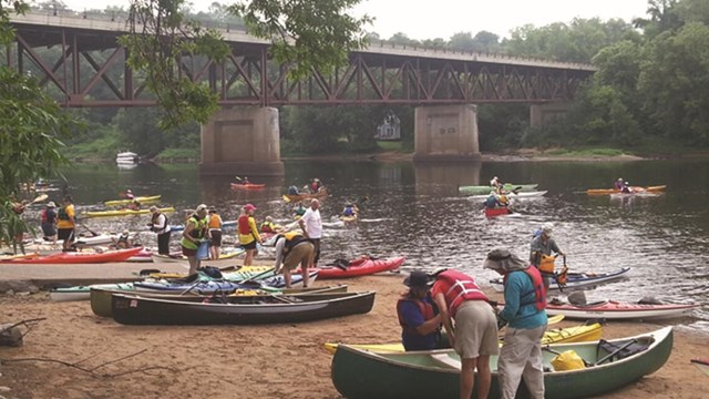 People milling around canoes and kayaks on the sandy landing while others approach from the river