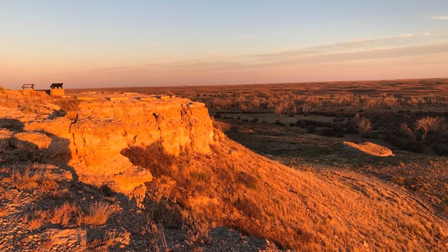 A rocky bluff overlooks the prarie, under a setting sun and warm light.