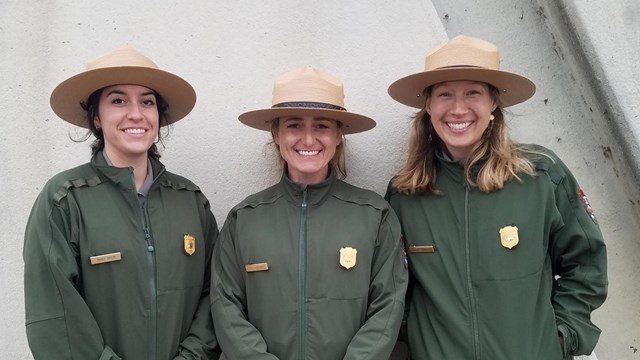 Three uniformed park rangers stand shoulder to shoulder.