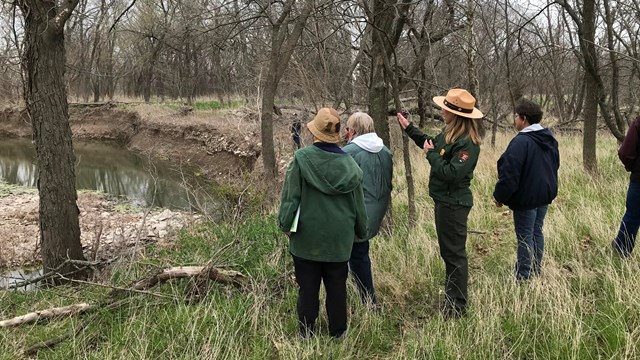 A uniformed park ranger stands with some members of the public, in a natural setting, pointing.