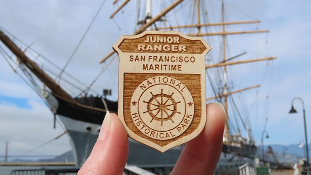 a San Francisco Maritime Junior Ranger Badge is held up, with a ship visible in the background.
